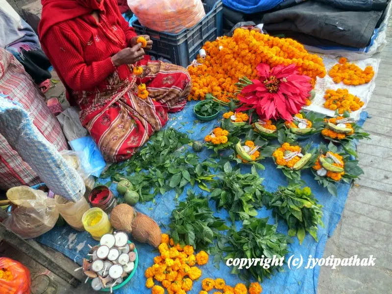 A flower vendor is busy making marigold flower garlands (सयपत्री फुल को माला) for deities and several other pooja items like belpatra leaves, dhaturo flowers, and fresh coconuts for sale in front of a Shiva Temple in Kathmandu.